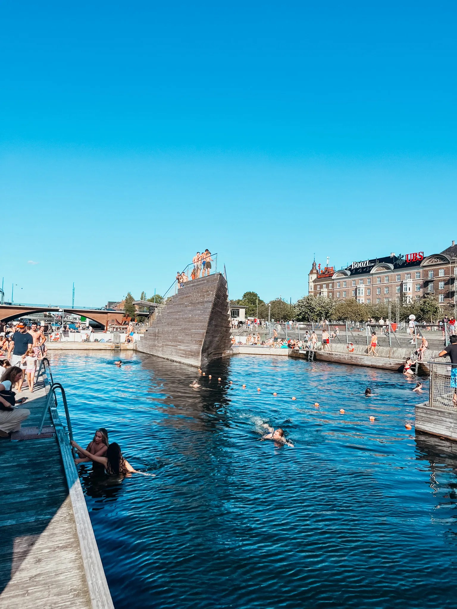People enjoying a swim in the harbour in Copenhagen. It's a bright blue sunny day. People are diving in the water off large wooden structures.