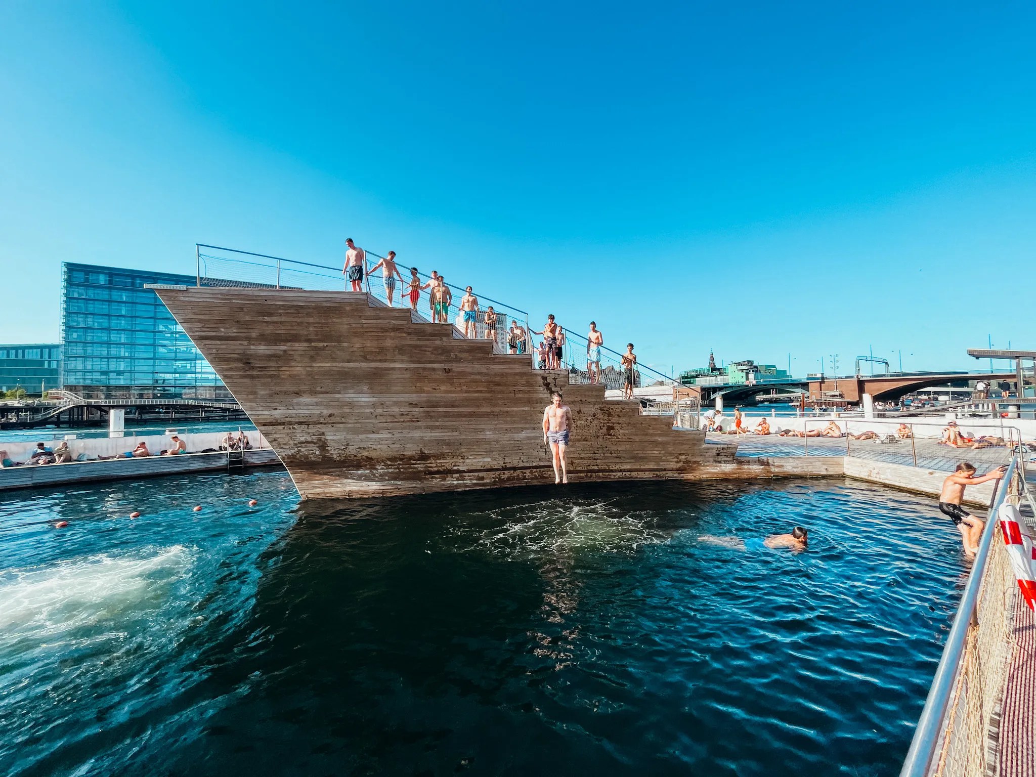 People enjoying a swim in the harbour in Copenhagen. It's a bright blue sunny day. People are diving in the water off large wooden structures.